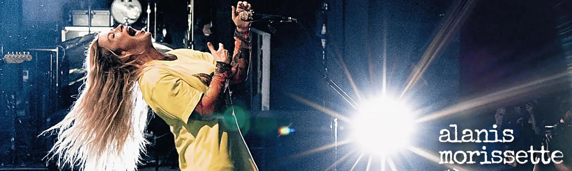 Alanis morissette performs on stage in a yellow shirt under bright spotlight.