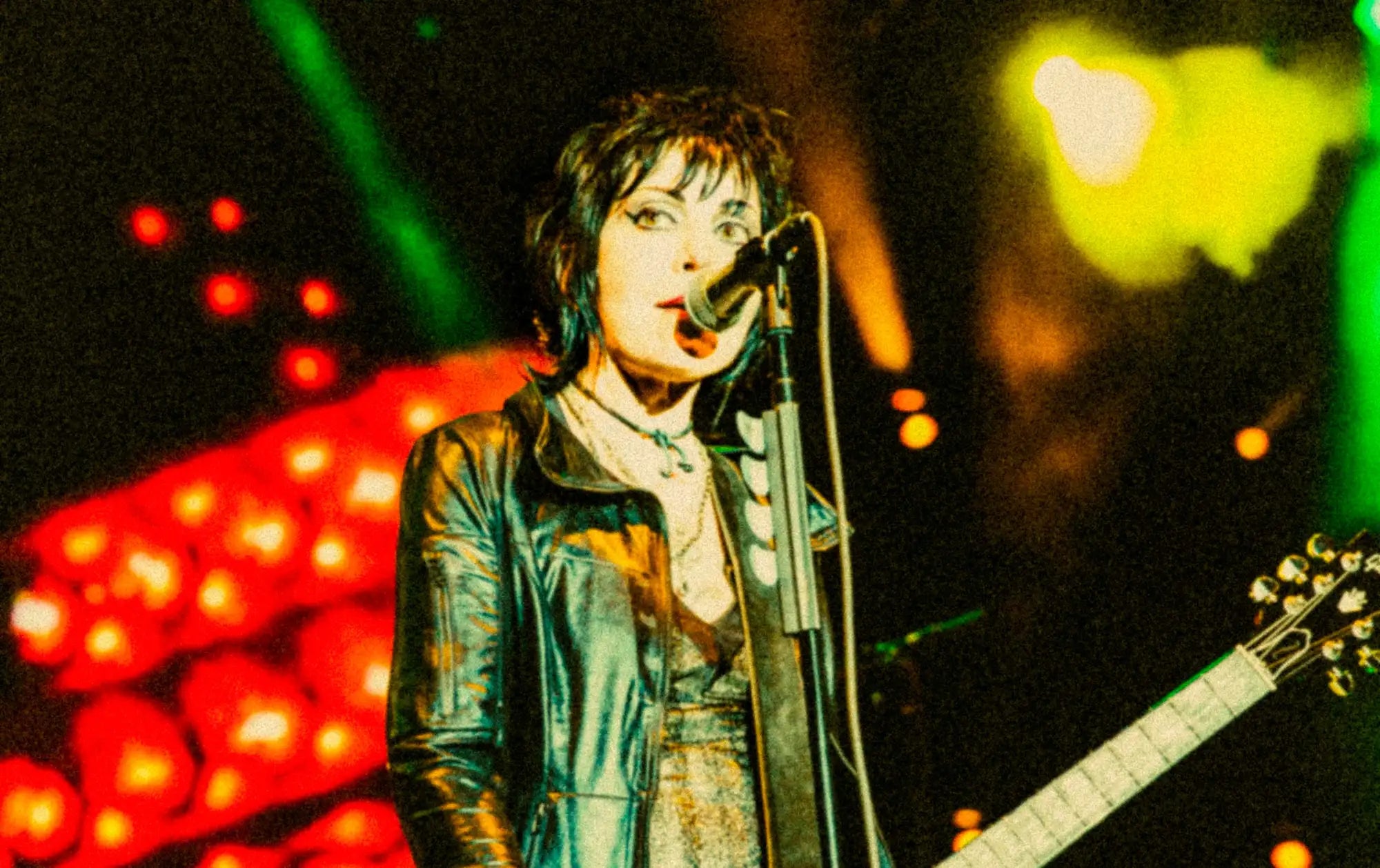A performer in a dark, glossy leather jacket sings into a microphone under stage lights.